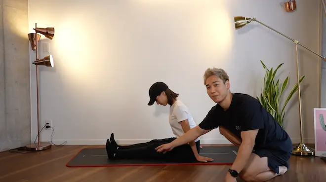 パーソナルトレーニングジムKARADA BESTAで下半身のむくみを取るストレッチを行う女性と指導するパーソナルトレーナー。女性はマットの上に足を伸ばして座り、背筋を伸ばしたまま上半身を前傾させて足をストレッチさせている。